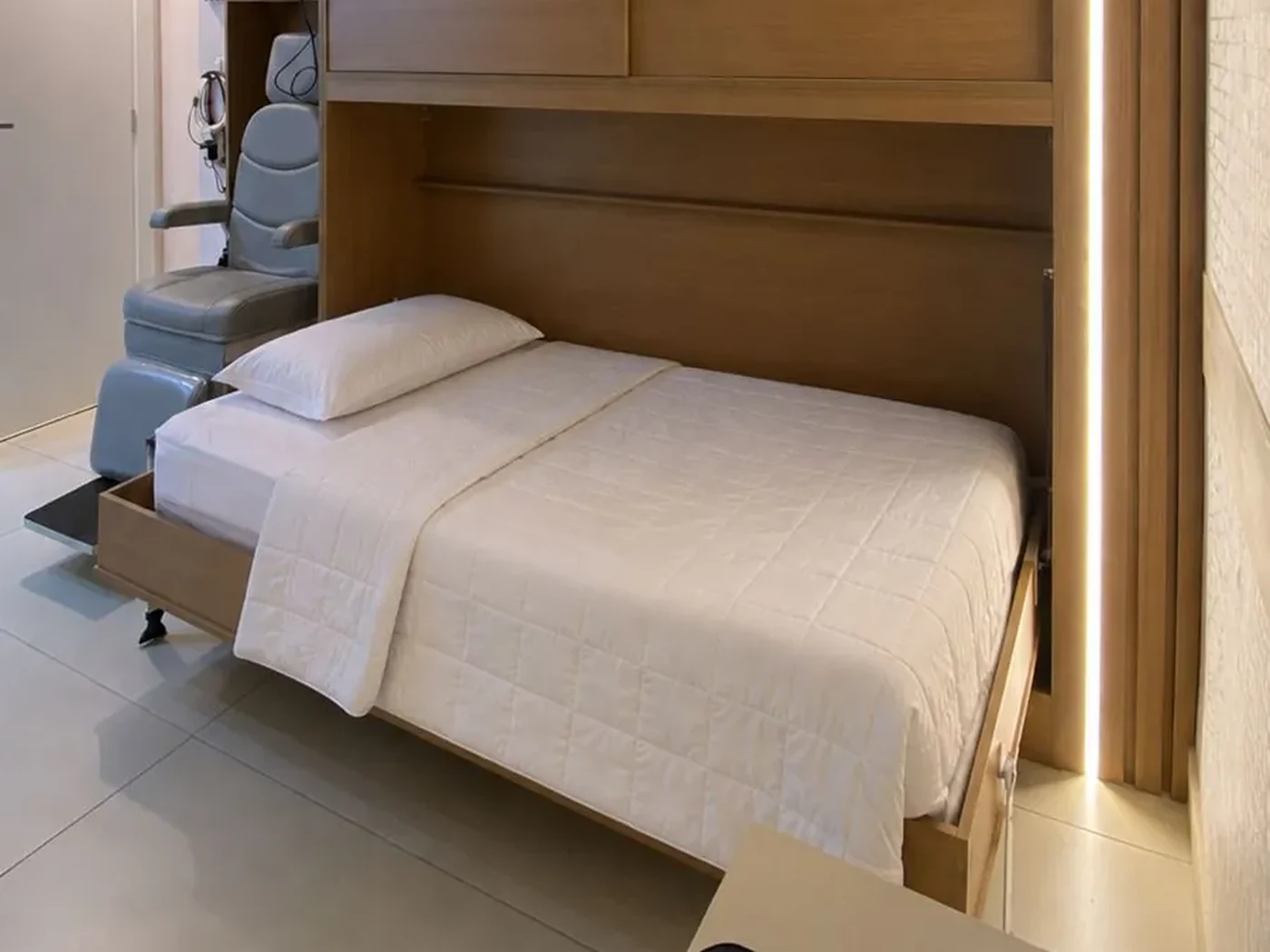 Quarto individual da MedSono em Taguatinga: cama de casal, marcenaria clara com iluminação indireta e poltrona ao lado